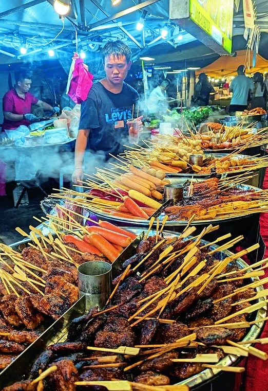 A vendor prepares skewers beneath the colourful lights of Taman Connaught market